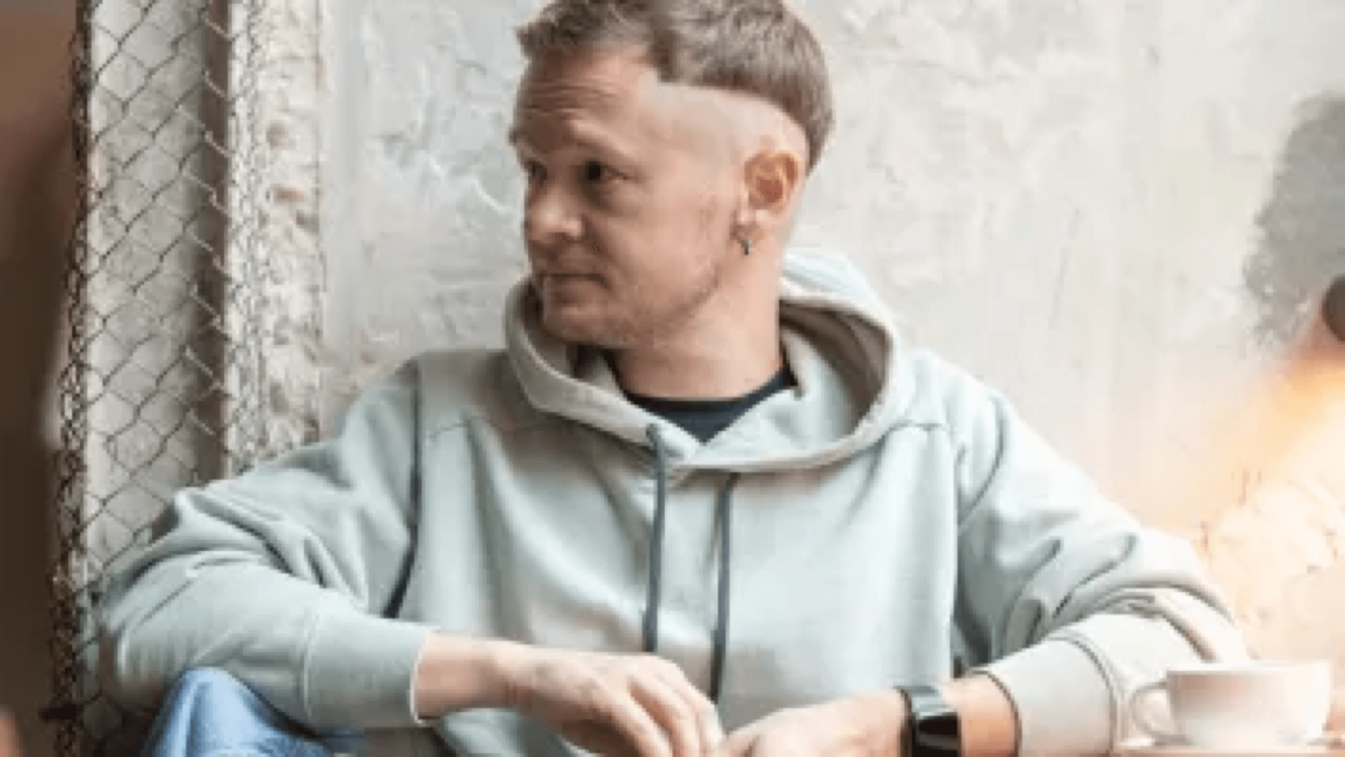 Man in a hoodie at a café table with a cup of coffee, looking thoughtfully aside in warm indoor light.
