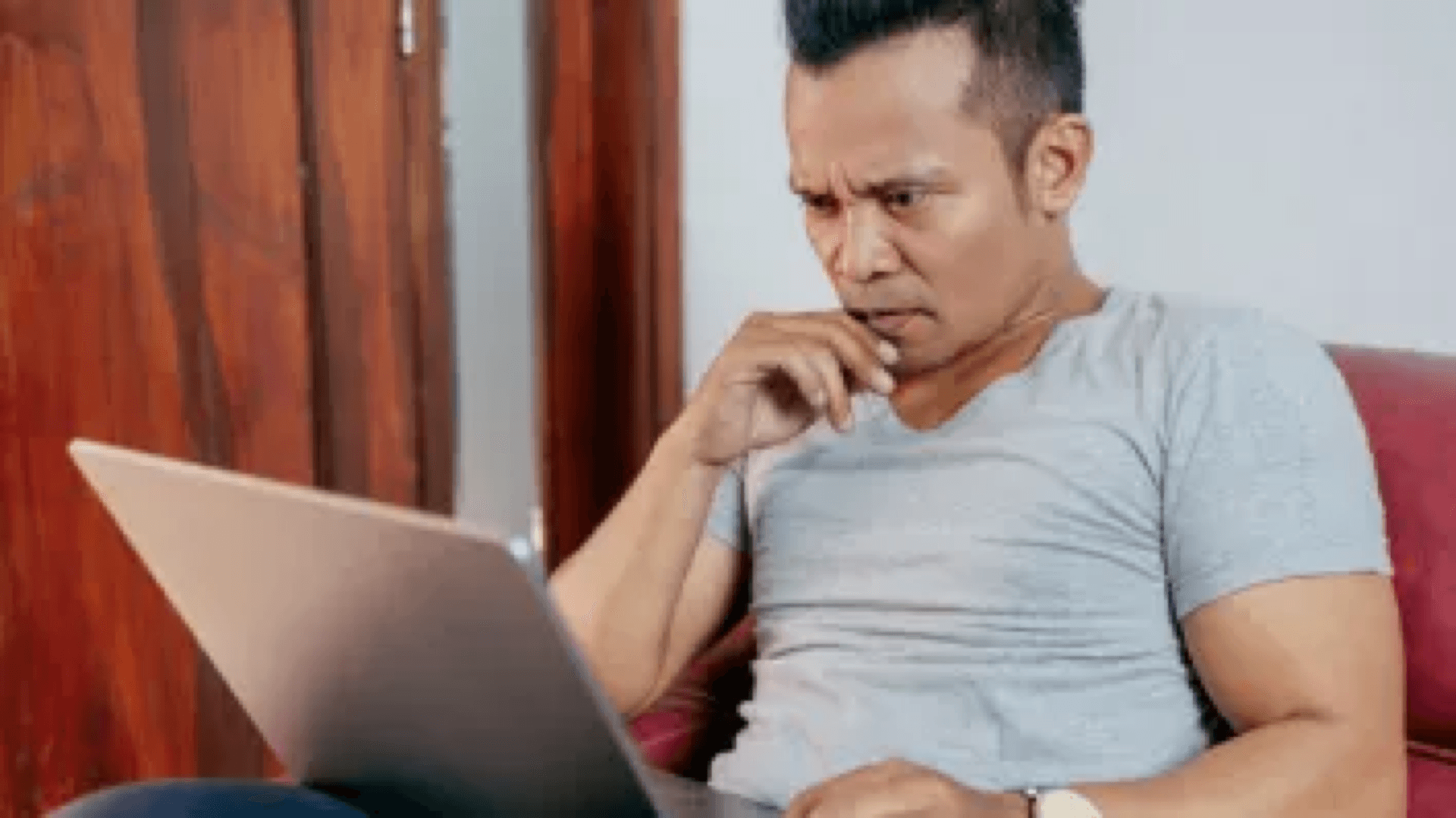 Man seated on a sofa focused on a laptop, hand to his mouth with a concerned expression.